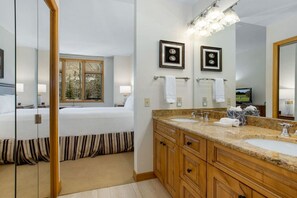 A hotel room with a bed, a large mirror, a vanity with a sink, and a window with a view of trees. - Highlands Lodge Beaver Creek - Corner Unit (Beaver Creek)