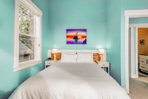 Quarto com uma cama grande, duas mesas de cabeceira com abajures, um quadro com um pôr do sol sobre a água e uma janela à esquerda.