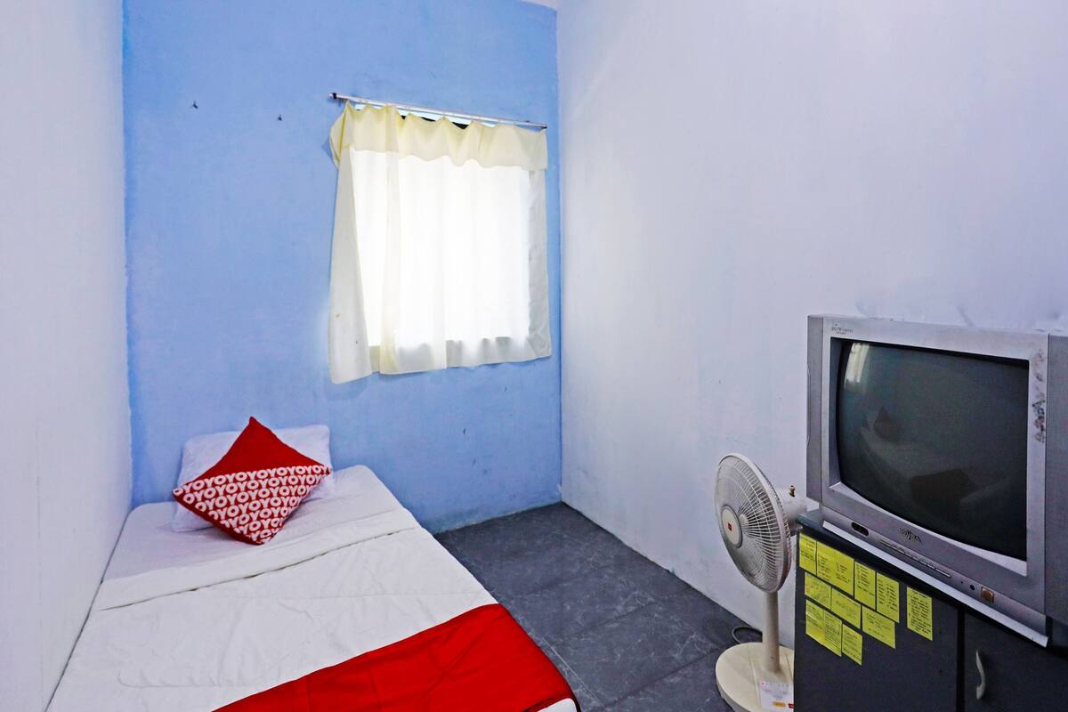 A small room with a bed, a window with a curtain, a television, and a fan.