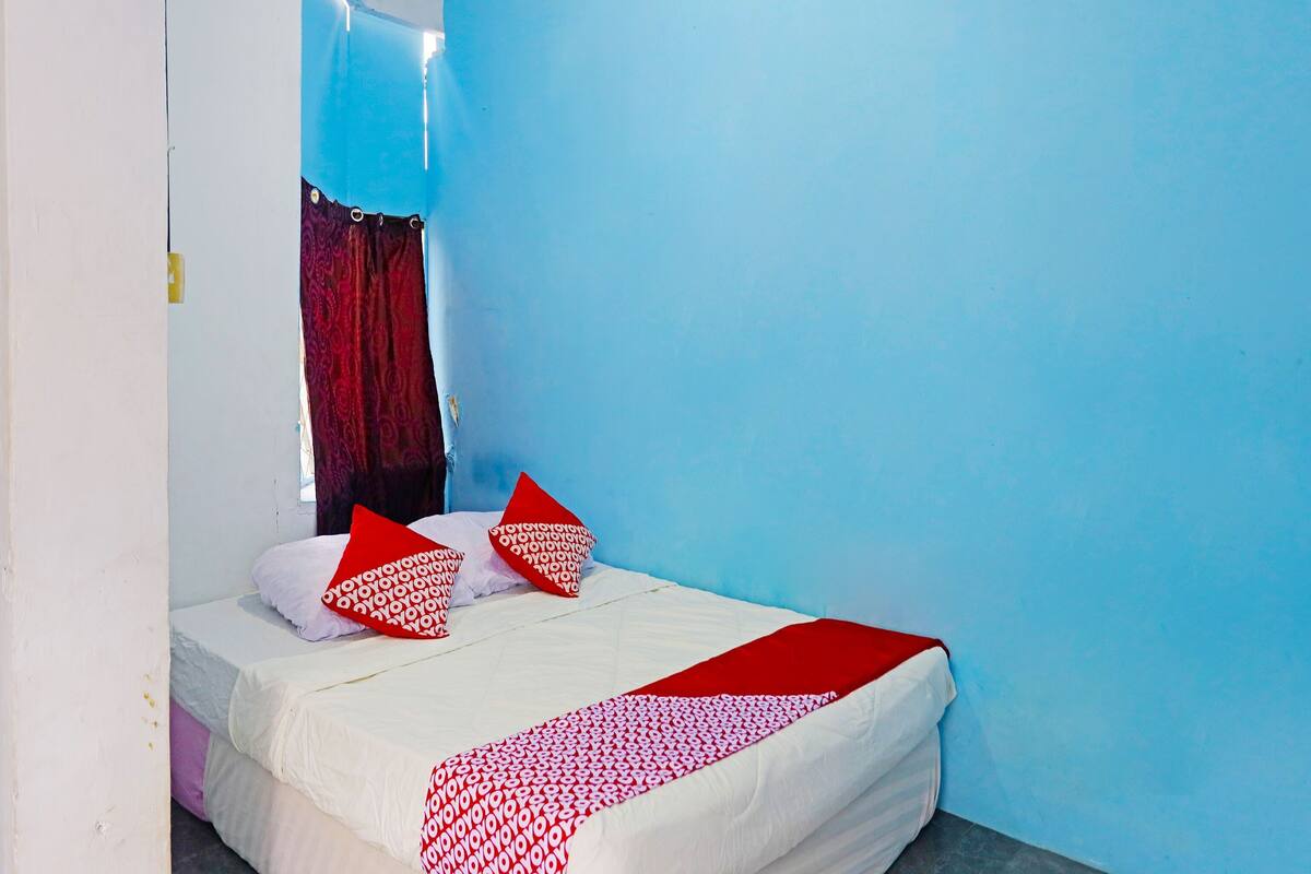 A single bed with white bedding and red pillows, a red and white patterned blanket, and a blue wall.