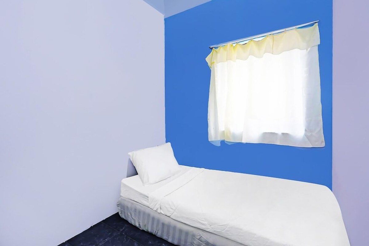 A small, single-bed room with a blue accent wall, a white bed, and a window with a yellow curtain.