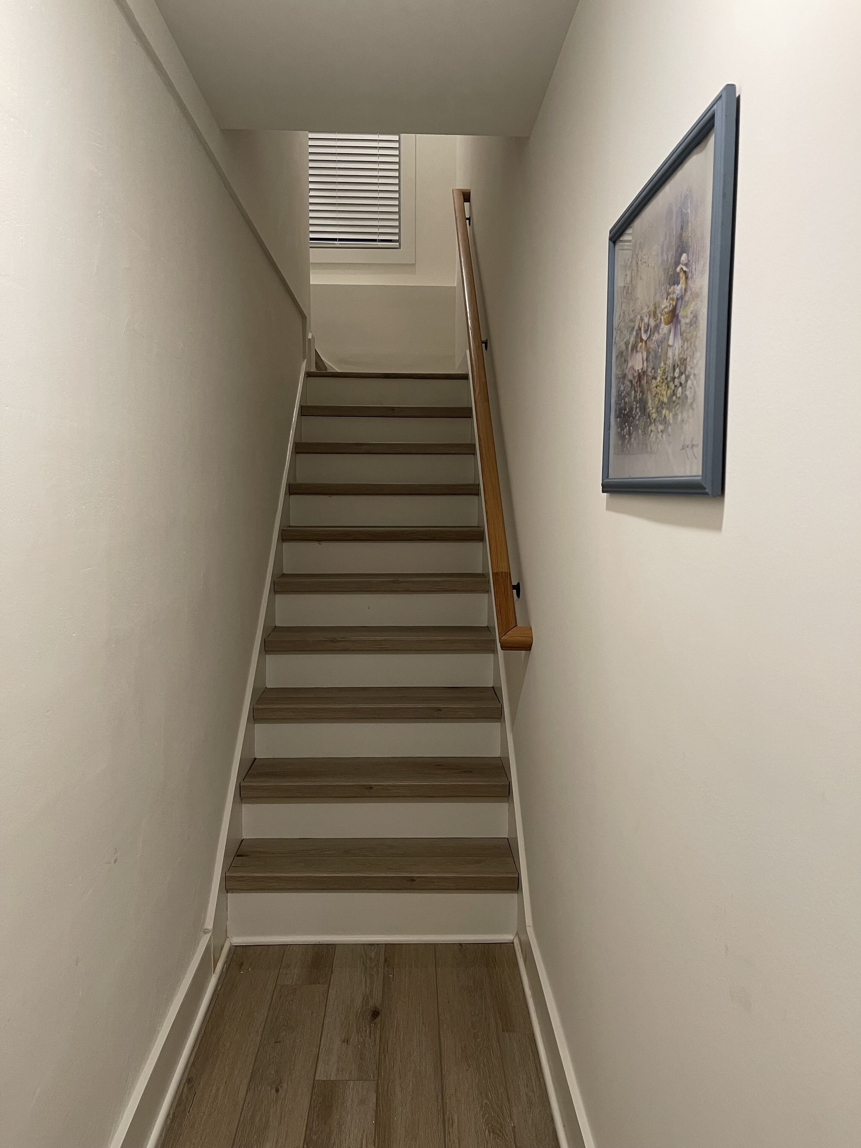 Un escalier avec des marches en bois et une rampe, menant à un palier doté d’une fenêtre et d’un tableau encadré au mur.