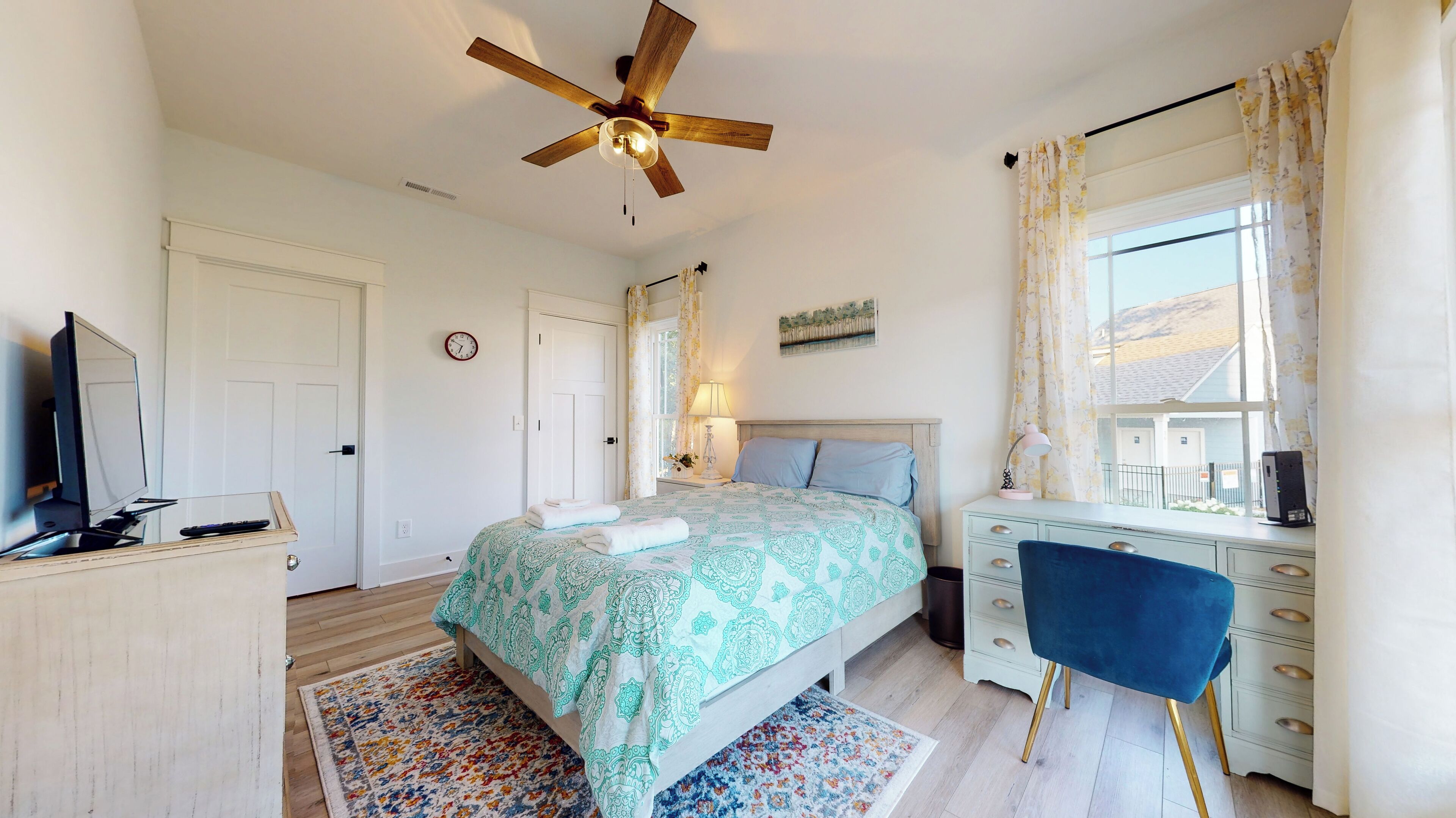 A bedroom with a bed, a desk, a chair, a ceiling fan, a window with curtains, and a TV on a stand.
