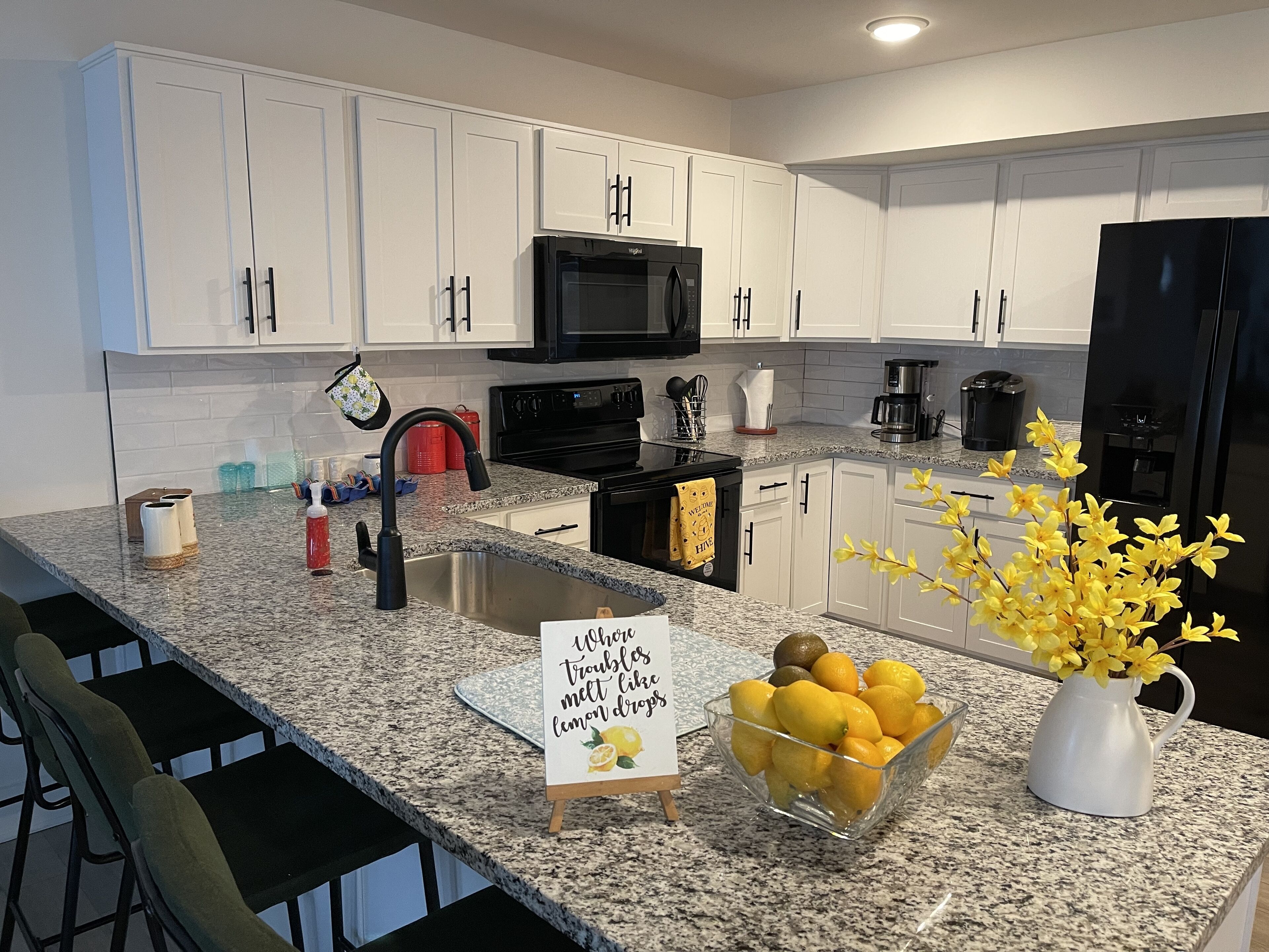 A modern kitchen with white cabinets, a granite countertop, a black refrigerator, and a bowl of lemons.