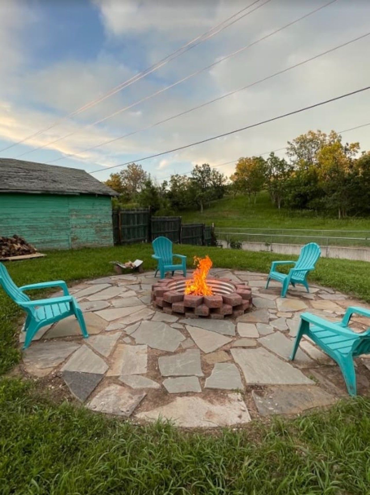 A circular stone fire pit with a fire, surrounded by four turquoise chairs, set on a stone patio with a grassy yard and a wooden shed in the background.