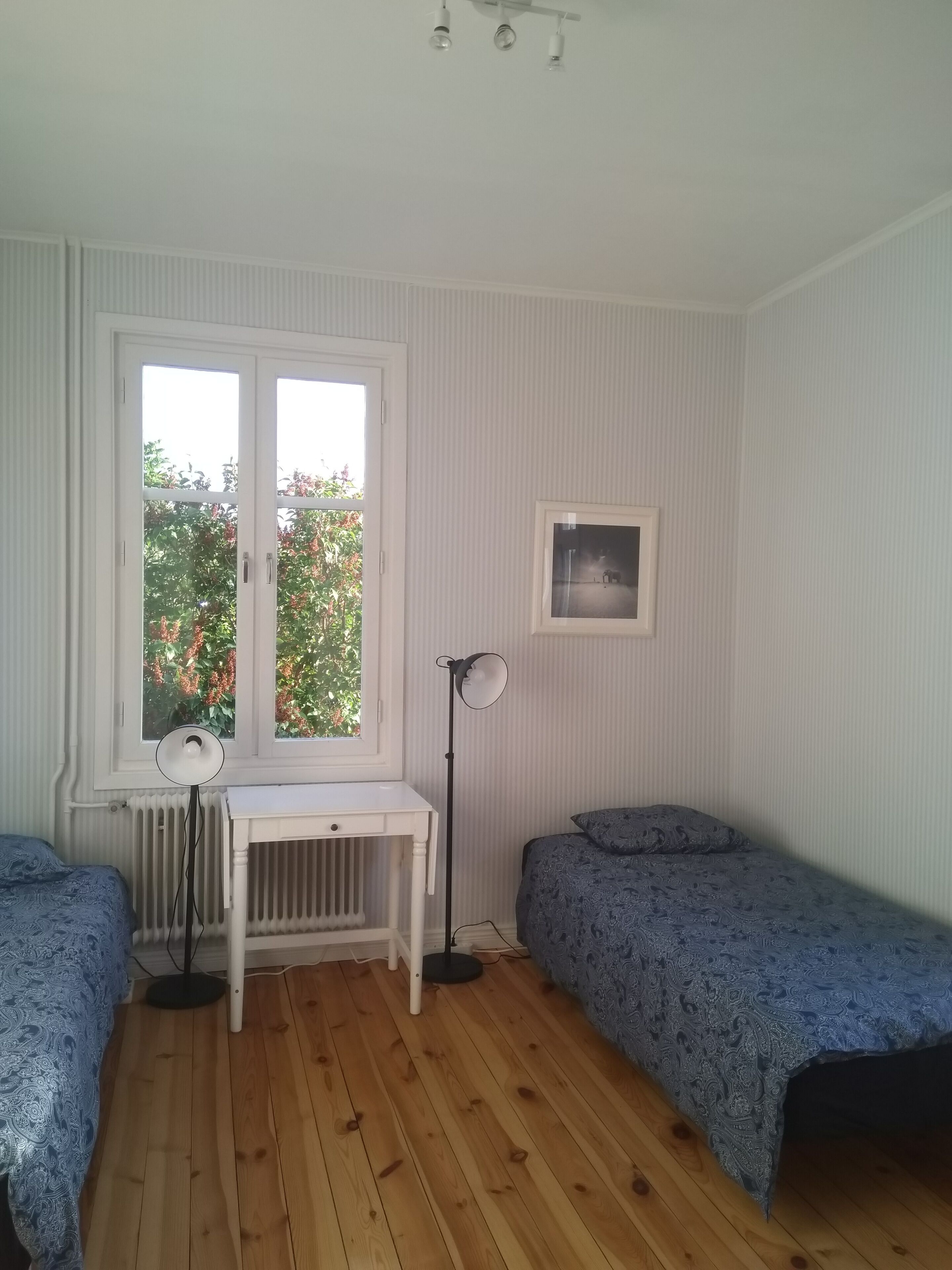 Una habitación con piso de madera, un radiador blanco, un escritorio con silla, dos camas individuales con ropa de cama estampada, una lámpara de pie y una ventana con vistas a la vegetación.