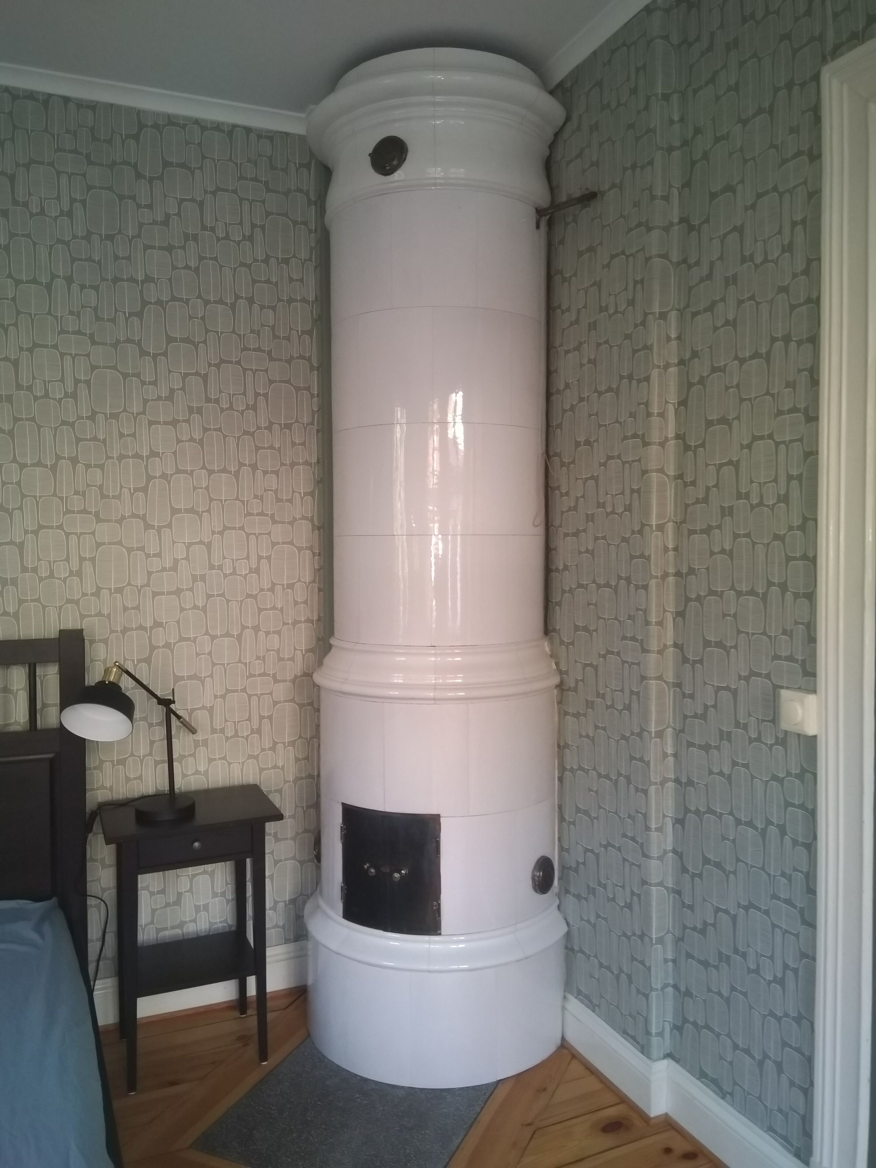 Una habitación con un calentador cilíndrico blanco, una mesita con una lámpara y un papel tapiz estampado.