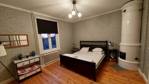Une chambre avec un plancher en bois, un lit avec une literie blanche, une fenêtre avec un store noir, un radiateur, une table de chevet avec une lampe, et un objet cylindrique blanc fixé au mur.