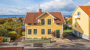 Une maison jaune à deux étages, avec un toit recouvert de tuiles rouges, des fenêtres vertes et un petit porche doté d’un banc.