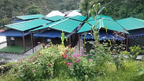 A row of rustic huts with green roofs, surrounded by lush greenery and flowering plants. - Phulwari Resort (Ukhimath)
