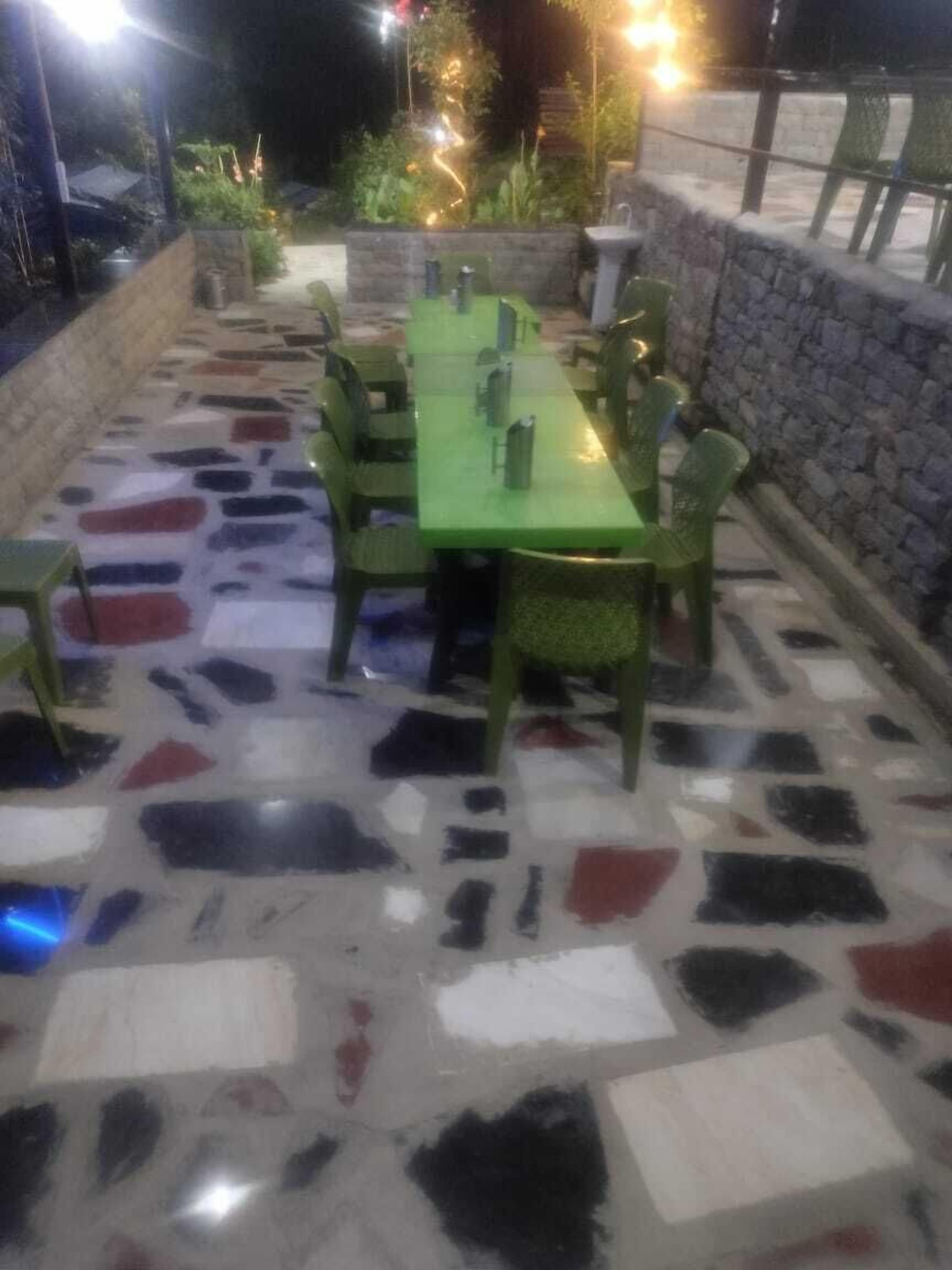 Un área de comedor al aire libre con una mesa y sillas verdes, rodeada de un muro de piedra y plantas.