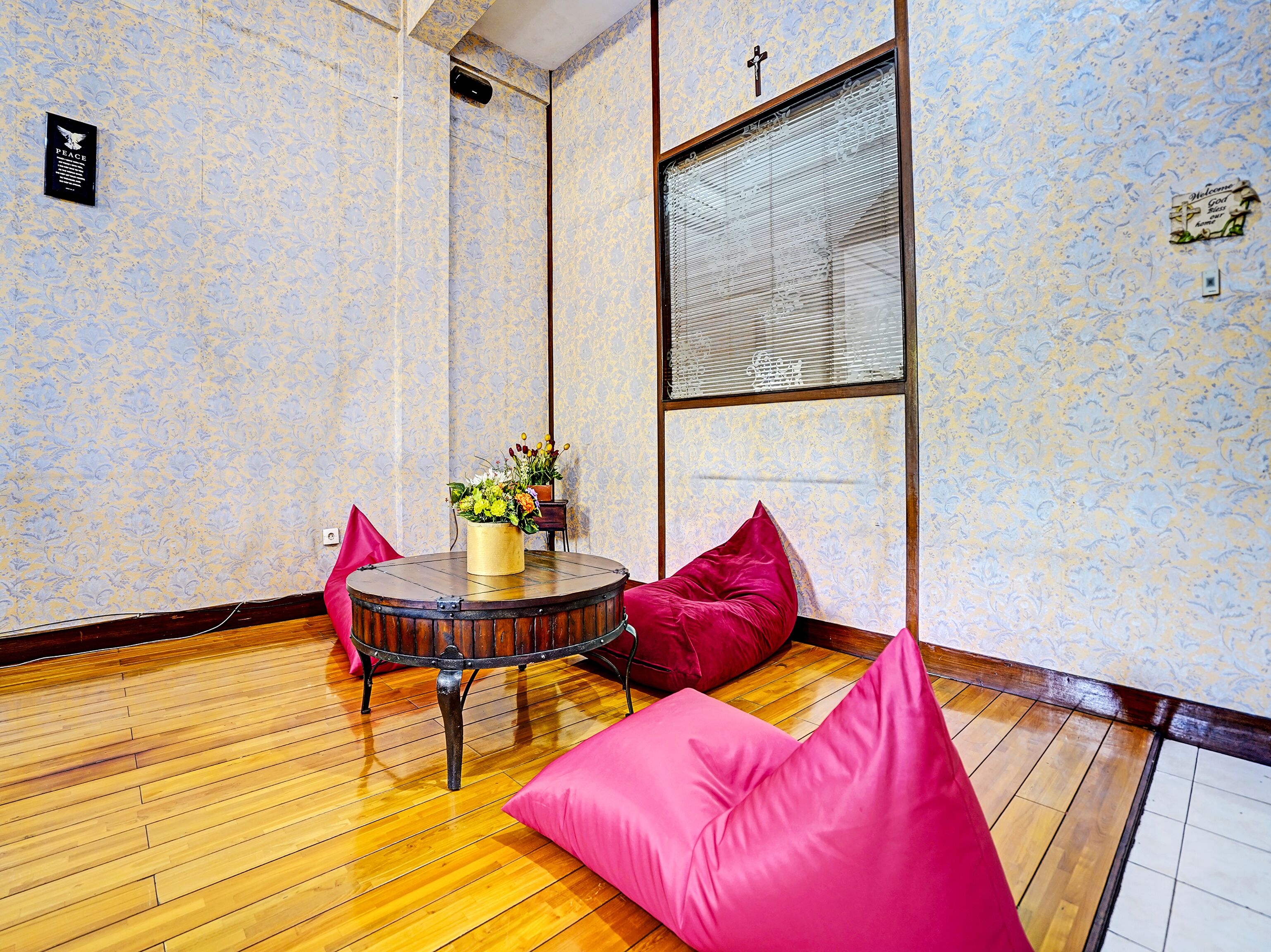 A room with patterned wallpaper, a wooden floor, a round table with a vase of flowers, and two pink cushions.