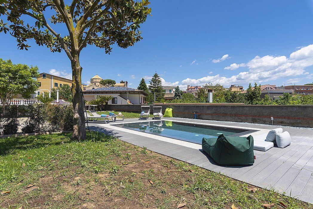 A swimming pool with lounge chairs and a green bean bag chair, surrounded by a grassy area and a tree.