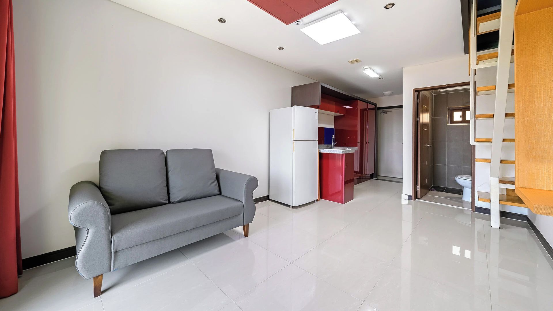 A modern living space with a grey sofa, a white refrigerator, and a red kitchen area.