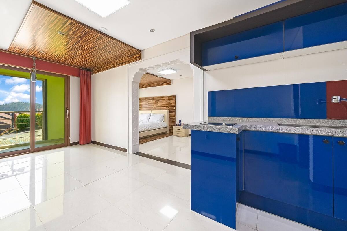 A modern hotel room with a large sliding glass door leading to a balcony, a separate bedroom with a wooden headboard, and a kitchen area with blue cabinets.