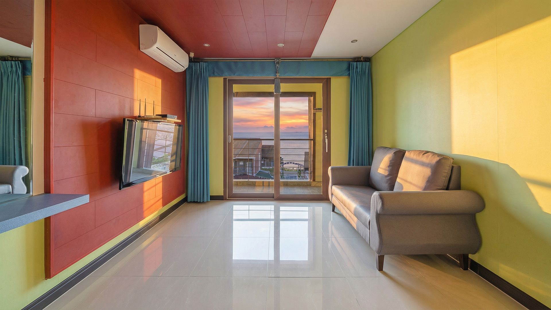 A modern hotel room with a flat-screen TV mounted on the wall, a sofa, and a sliding glass door leading to a balcony with a sunset view.