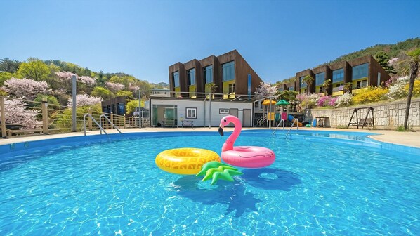 A swimming pool with inflatable flamingo and starfish floats, surrounded by a wooden fence and a building with large windows. - Geojedo Pilos Ocean View Pension (Geoje)