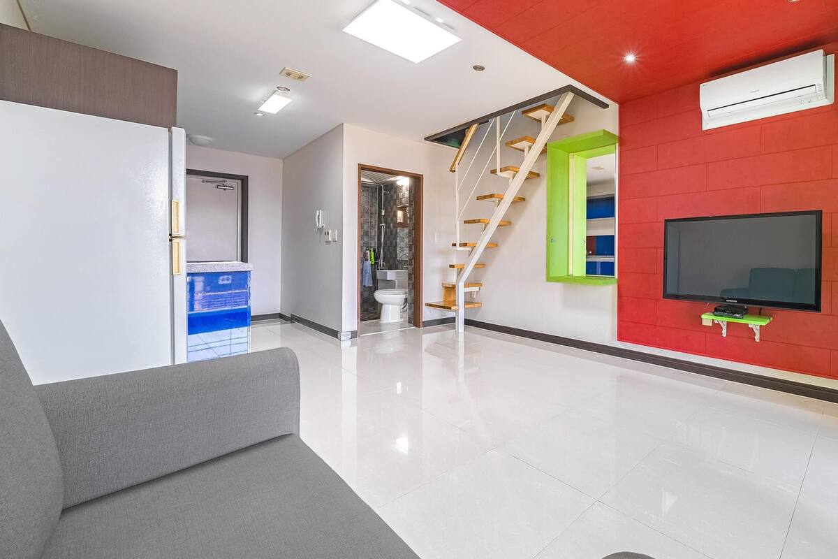 A modern living room with a flat-screen TV mounted on a red wall, a staircase with wooden steps, and a grey sofa.