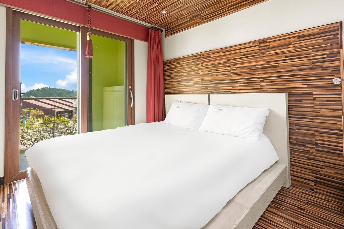 A bedroom with a large bed, wooden panel wall, and a sliding glass door leading to a balcony with a view of greenery.
