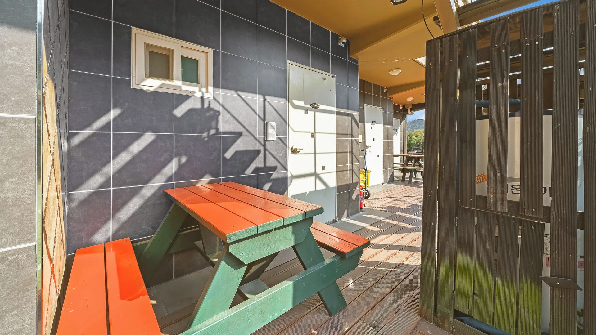 A patio area with a wooden table and benches, a door with a keypad lock, and a tiled wall with a window.