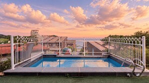 Une piscine entourée d’une clôture blanche, avec de la verdure tout autour et des bâtiments en arrière-plan.