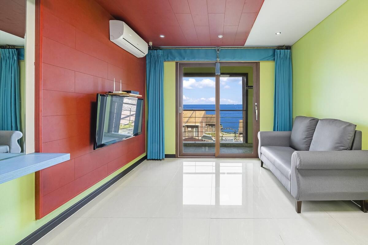 A modern living room with a grey sofa, a flat-screen TV mounted on the wall, and a balcony with a view of the sea.