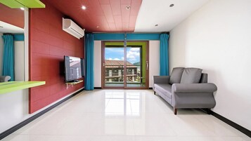 Una sala de estar moderna con un televisor de pantalla plana montado en una pared roja, un sofá gris y un puerta corrediza de vidrio que da a un balcón con vistas a edificios.