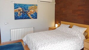 Ein Schlafzimmer mit einem hölzernen Kopfteil, einer weißen floralen Bettdecke, einem blauen Teppich, einem Gemälde einer Meerjungfrau und einem Deckenventilator.