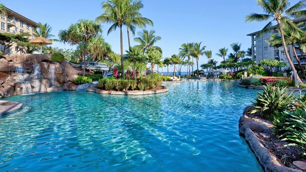Ein Resort-Pool mit klarem, blauem Wasser, Palmen und einer Wasserfall-Anlage aus Felsen.