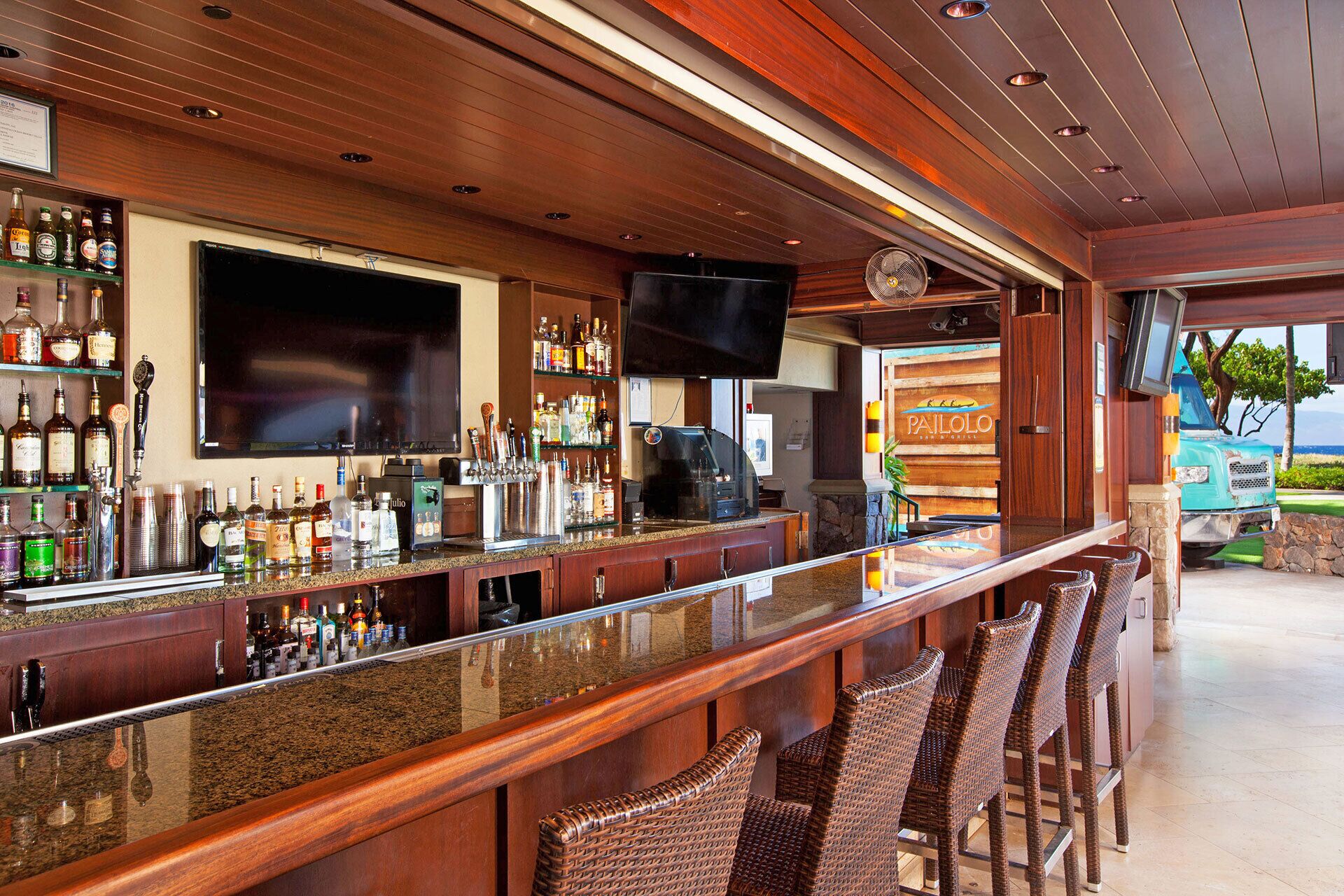 A bar area with a long counter, multiple bottles, a television, and wicker chairs.