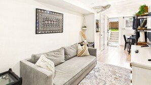 Una sala moderna con un sofá gris, un tapiz decorativo en la pared y una zona de comedor con mesa y sillas.