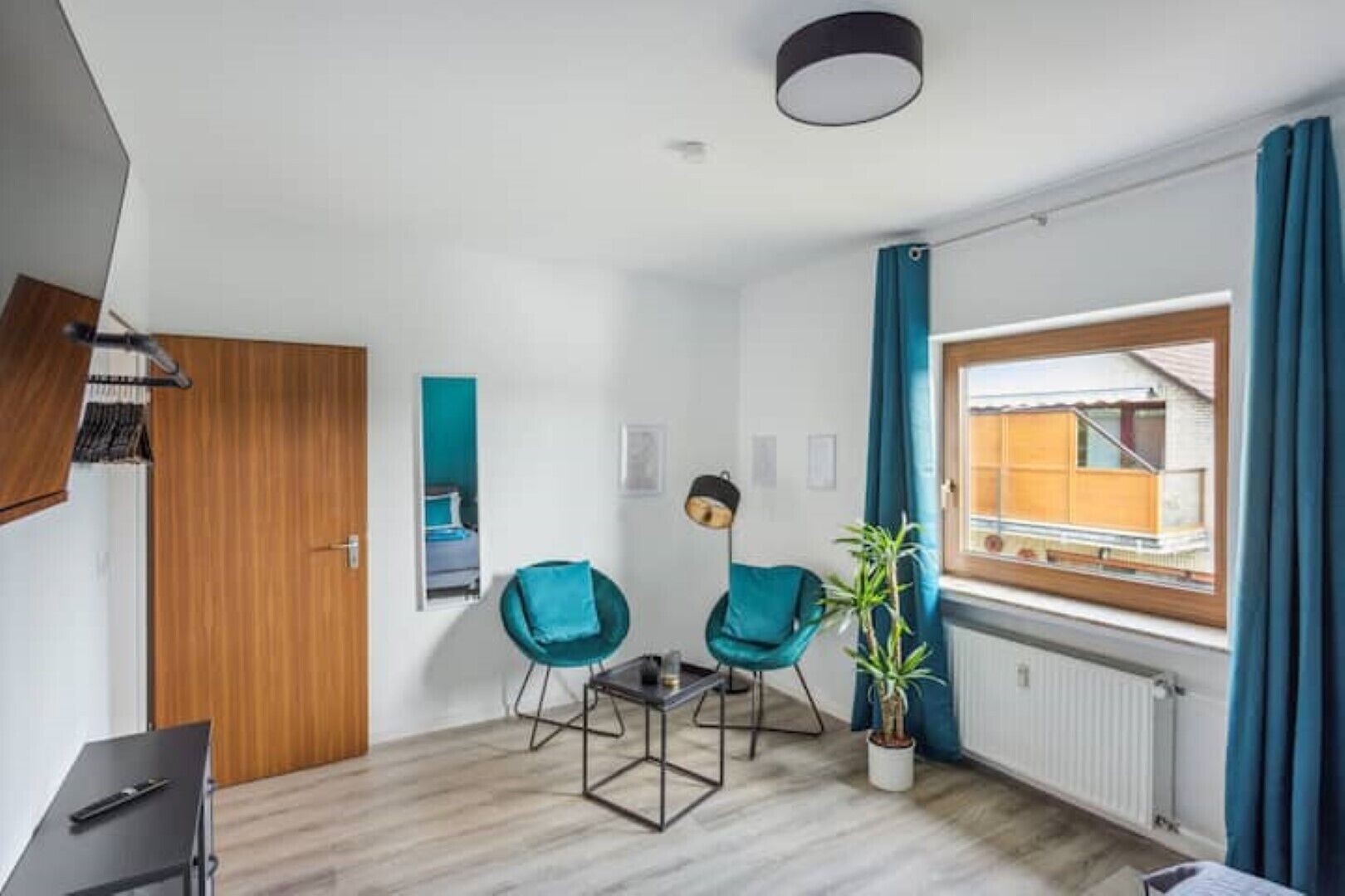 A modern room with a wooden door, a small table, two teal chairs, a floor lamp, a potted plant, and a window with curtains.