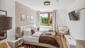 Moderni hotellihuone, jossa on suuri sänky, työpöytä, tuoli, televisio ja ikkuna, josta avautuu näkymä vehreään maisemaan.
