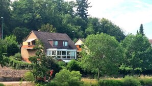 Une maison dotée d’une grande baie vitrée, entourée d’une végétation luxuriante et bordée par une rivière.