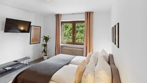 Une chambre à coucher avec un lit, une télévision, une fenêtre avec des rideaux, une plante et des tableaux encadrés accrochés au mur.