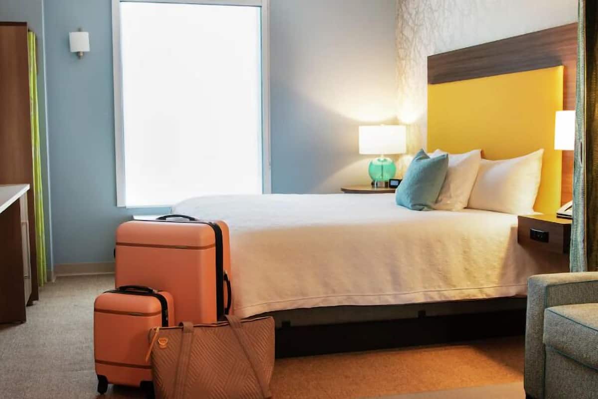 A hotel room with a bed, a nightstand with a lamp, a suitcase, and a chair.