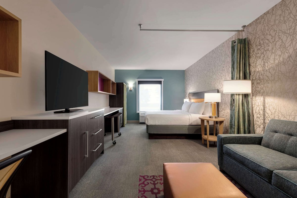 A hotel room with a bed, a desk with a flat-screen TV, a sofa, a lamp, and a window with curtains.