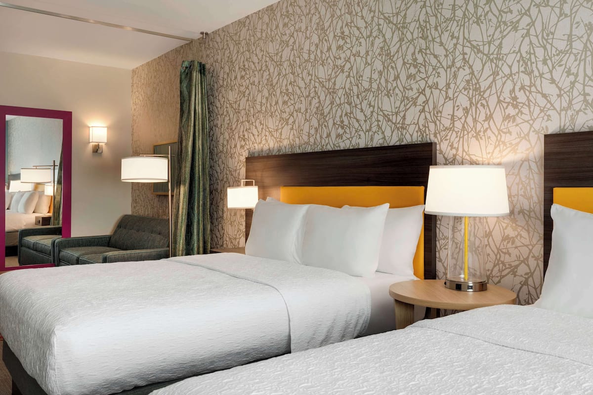 A hotel room with two beds, a sitting area with a chair, a round table, and a mirror.