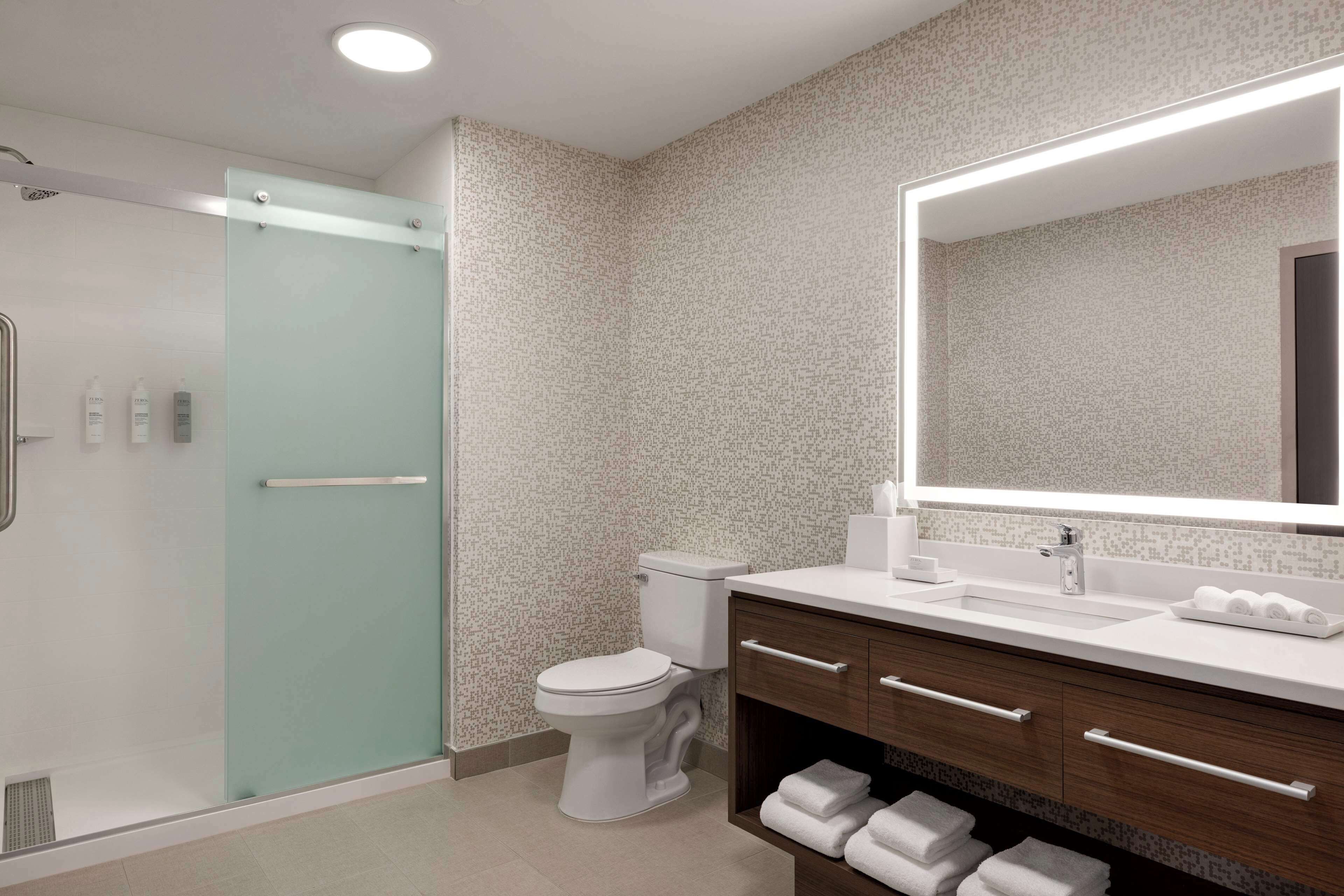 A modern bathroom with a glass shower enclosure, a white toilet, a large mirror, and a wooden vanity with towels.