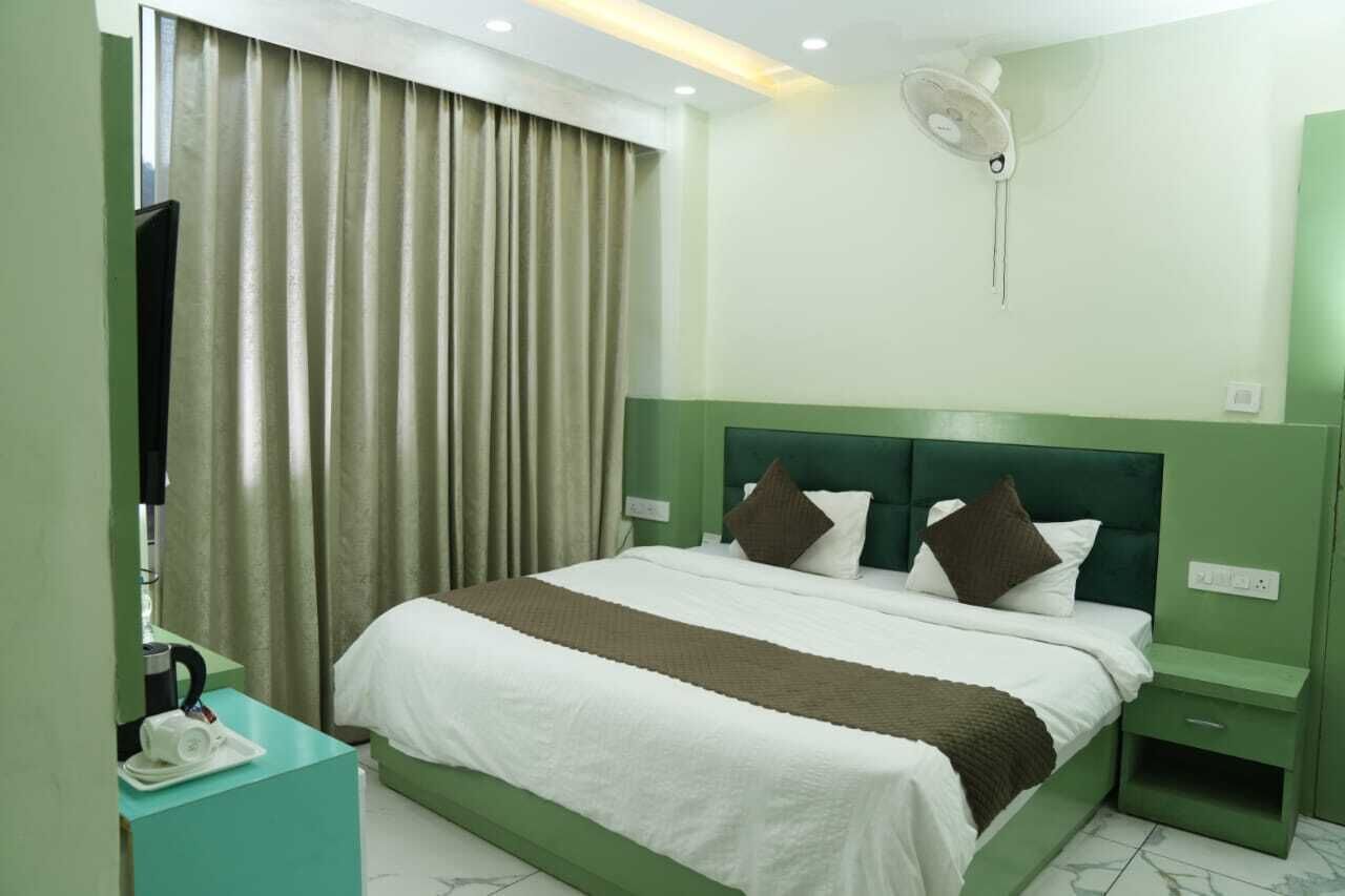A hotel room with a bed, a nightstand, a desk with a phone and a kettle, a window with curtains, and a wall-mounted fan.