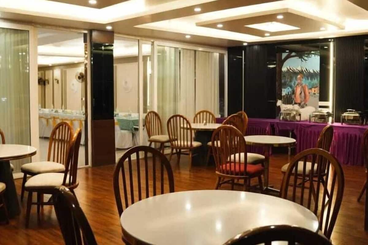 Un restaurante con mesas y sillas de madera, un escenario con un telón de fondo y una zona de cocina visible a través de un panel de vidrio.
