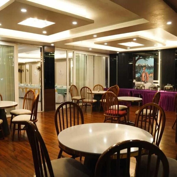 A restaurant with wooden tables and chairs, a stage with a backdrop, and a kitchen area visible through a glass partition.