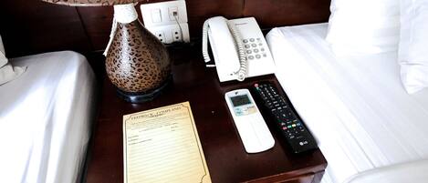 Una cama de hotel bien tendida con una mesita de noche que contiene una lámpara, un teléfono, un control remoto y un formulario de comentarios.