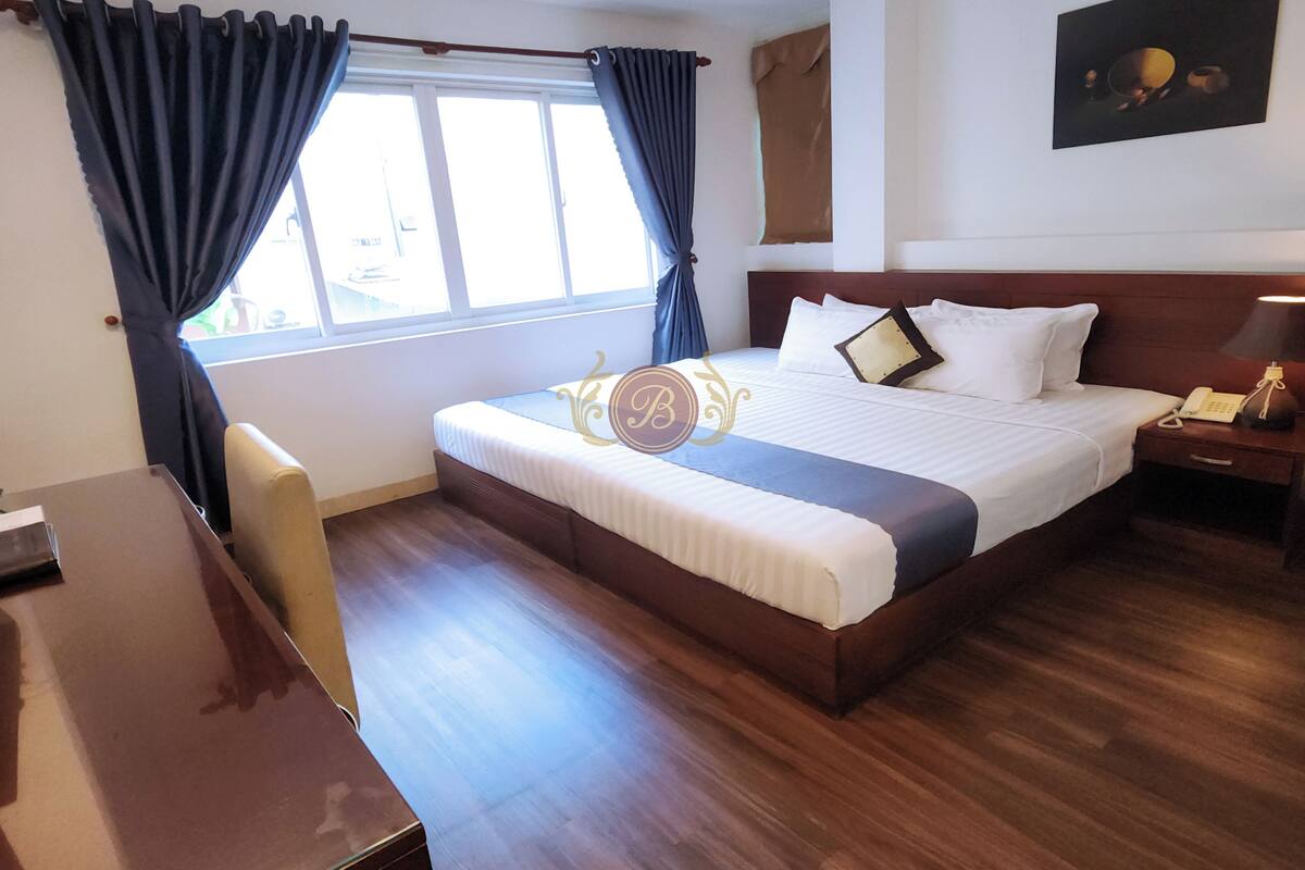 A hotel room with a large bed, a desk, and a chair. There is a window with curtains, a painting on the wall, and a nightstand with a lamp.