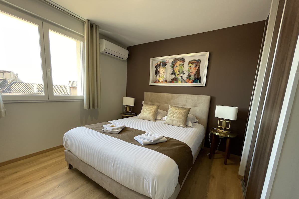 A hotel room with a large bed, two bedside tables with lamps, a painting on the wall, and an air conditioning unit.
