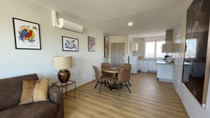 Eine moderne Wohnung mit Wohnzimmer, Essbereich und Küche. An den Wänden befinden sich gerahmte Kunstwerke, ein Sofa mit Kissen, ein Tisch mit Stühlen und eine Lampe.