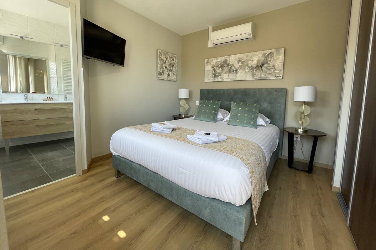 A bedroom with a bed, a TV, a wall-mounted air conditioner, and a bathroom visible through an open door.