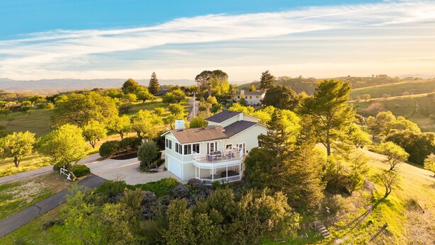 A large, two-story house with a balcony, surrounded by lush greenery and trees.