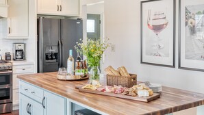 A kitchen with white cabinets, a stainless steel refrigerator, a wooden countertop, and a dining area with a basket of bread and a cutting board with cheese and meats.
