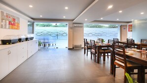A modern dining area with wooden tables and chairs, a buffet line with covered food containers, and a view of a green outdoor area through a glass partition. - Limewood Stay - Cyber Hub (Gurugram)